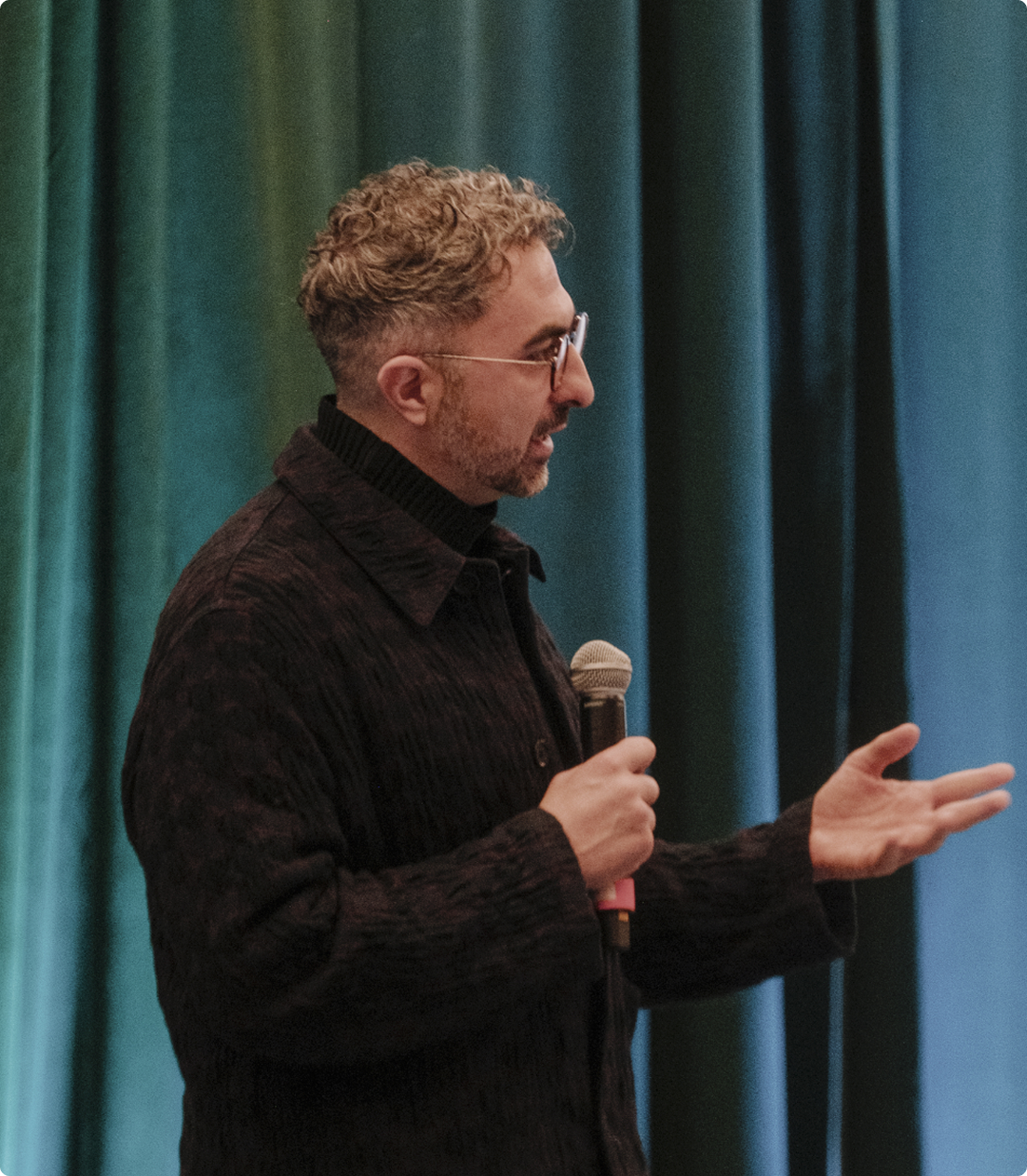 A man with short curly hair and glasses, wearing a dark sweater, speaks into a microphone while gesturing with his hand. He stands in front of a backdrop of blue and green curtains.